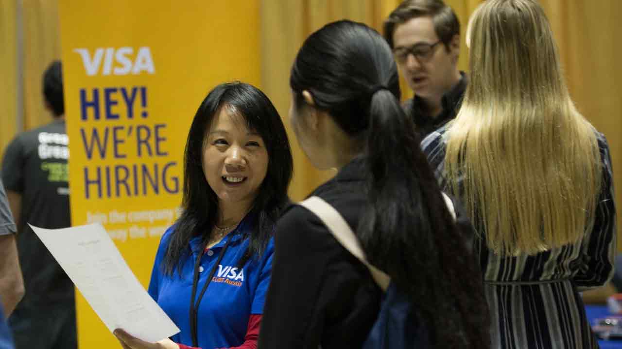 An employer for Visa meets with a student at a career fair while other students stand nearby a sign that says Visa: Hey! We're Hiring